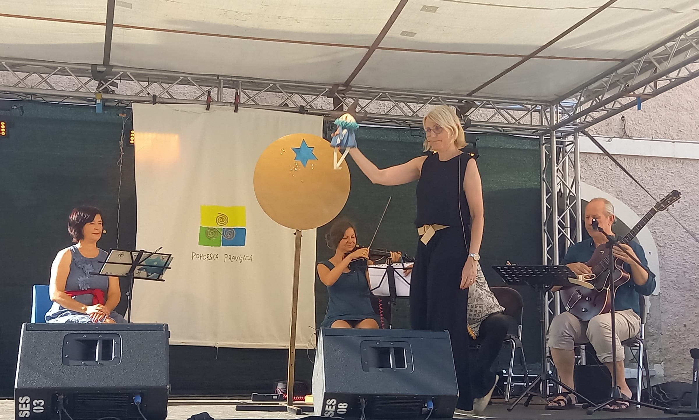 Pripovedovalka z lutko Agice na odru Pohorske pravljice, ob njej glasbeniki, Ruše, 19. 8. 2026