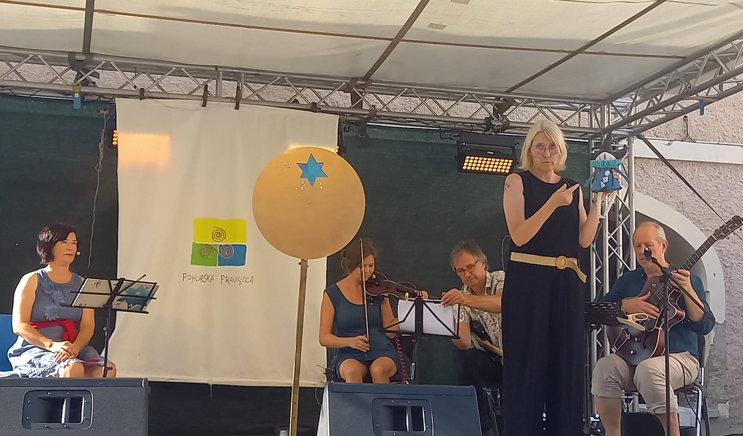 Glasbena pravljica Agica, mala čarovnica: pripovedovalka in glasbena zasedba na odru pred občino Ruše, 19. 8. 2026