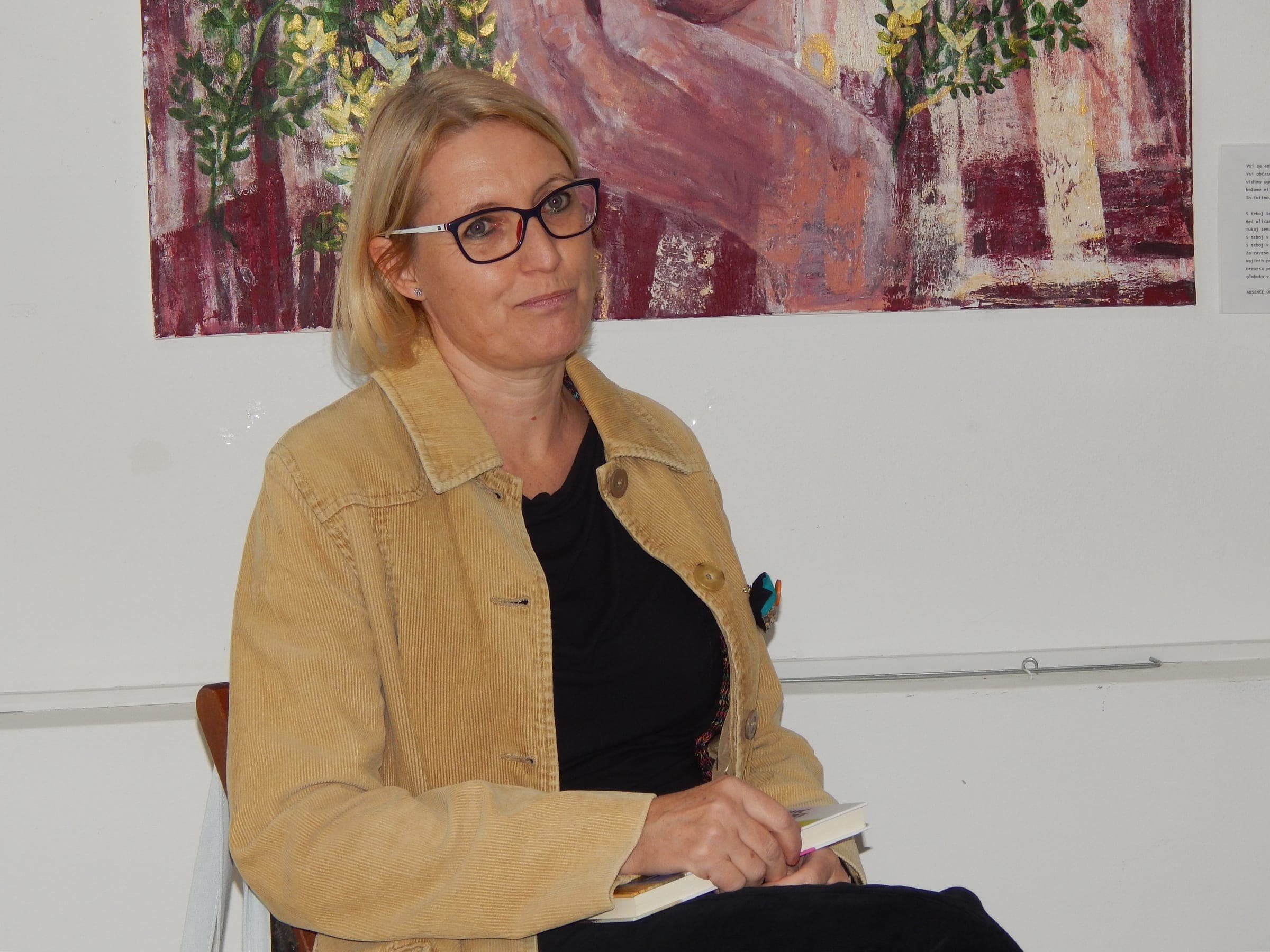 Sogovornica na predstavitvi romana Kavč učiteljice Veronike sedi pred sliko na steni, Literarna hiša Maribor, 2023